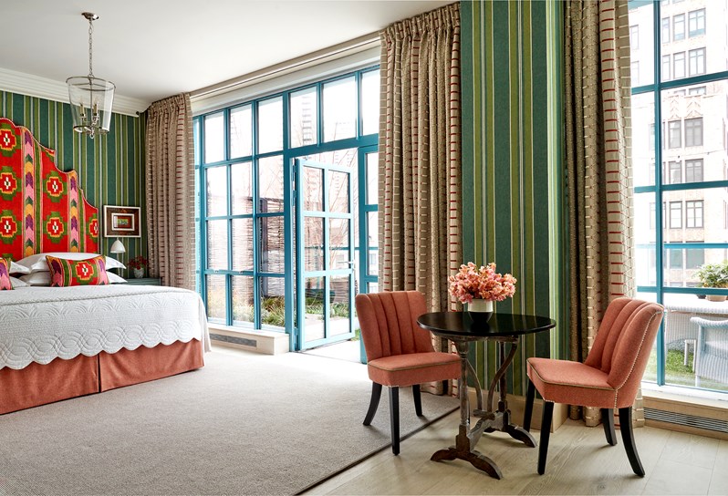 A bedroom with green striped walls and floor to ceiling windows and doors that open out onto a terrace. A bed with a multi-coloured geometric print headboard and matching cushions. A small round table by the window with two pink chairs either side.