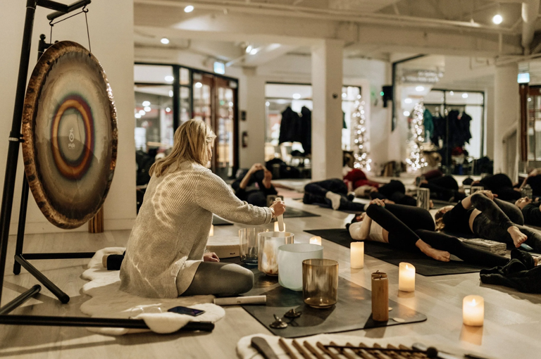 Sound practitioner Nancy Truman leading a sound bath class. Rows of women on yoga mats lie in front of her. She is playing glass sound bowls in front of her and there is a large gong behind her