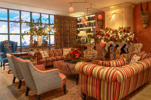 A diagonal shot of the drawing room at Warren Street Hotel. Two sofas and four arm chairs are placed around a coffee table, with a fireplace dressed with a festive garland and stockings. On the far side of the room, there is a table featuring a festive floral display, in front of windows looking through to the Orangery