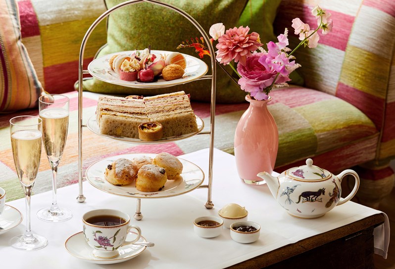 A three-tiered Summer themed afternoon tea. The first tier contains desserts such as a cherry-shaped cake, macaroons, etc. The second layer contains savoury bites. There are eight finger sandwiches as well as bite-sized quiches. The bottom tier contains four freshly baked scones with clotted cream and jam on the side. Accompanying on the table, there are 2 glasses of champagne as well as two English teas.