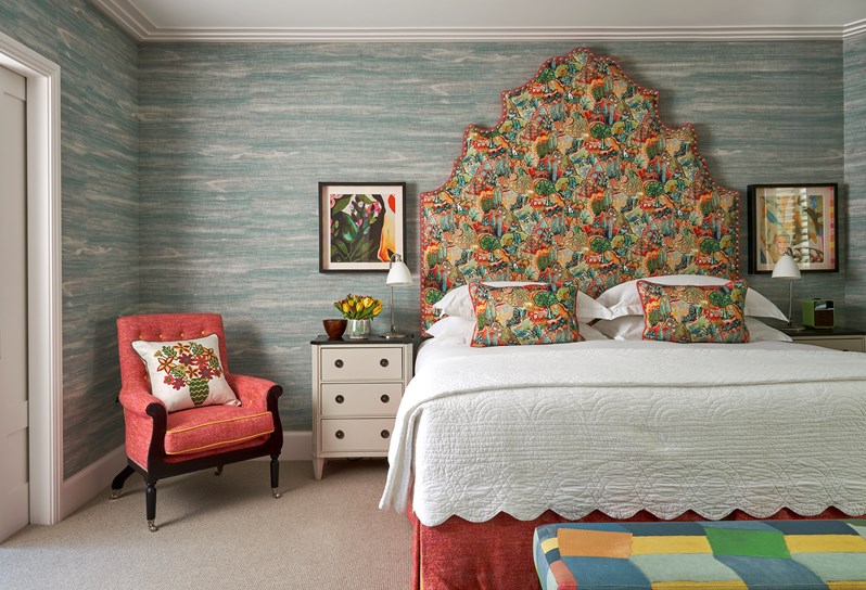 A front on image of the bedroom of The Deluxe Two Bedroom Suite, Room 5. A stylish bedroom featuring a vibrant patterned headboard and pillows, blue textured wallpaper, eclectic artwork, and an arm chair in the room.