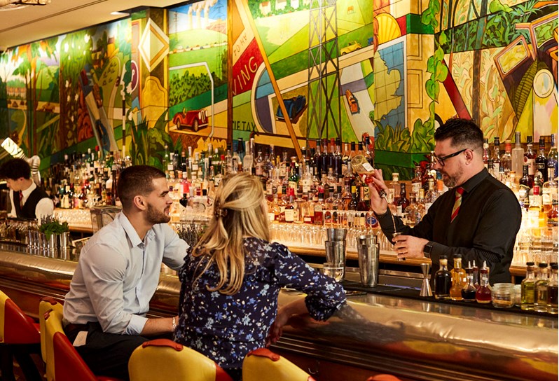 A man and a woman sat on high chairs at a bar. Behind the bar a male bartender is pouring a bottle of liqueur into a shot measurer, he is standing in front of multiple bottles of alcohol lined across the back of the wall.