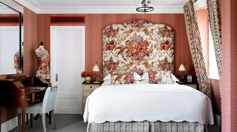 Salmon coloured bedroom with beautiful flower headboard, work space with desk and chair on the left side of the bed and widows on the left.