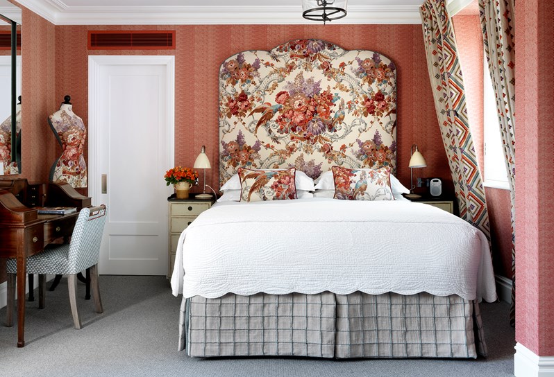 Salmon coloured bedroom with beautiful flower headboard, work space with desk and chair on the left side of the bed and widows on the left.