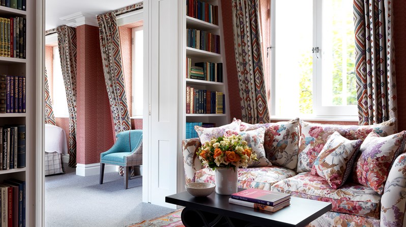 Living room of the Terrace Suite at Covent Garden Hotel with a flower pattern sofa and coffee table sitting over a patterned rug. To the left there is 2 bookcases with books on the wall, the door leading to the bedroom in between, where there is a chair and the corner of the bed