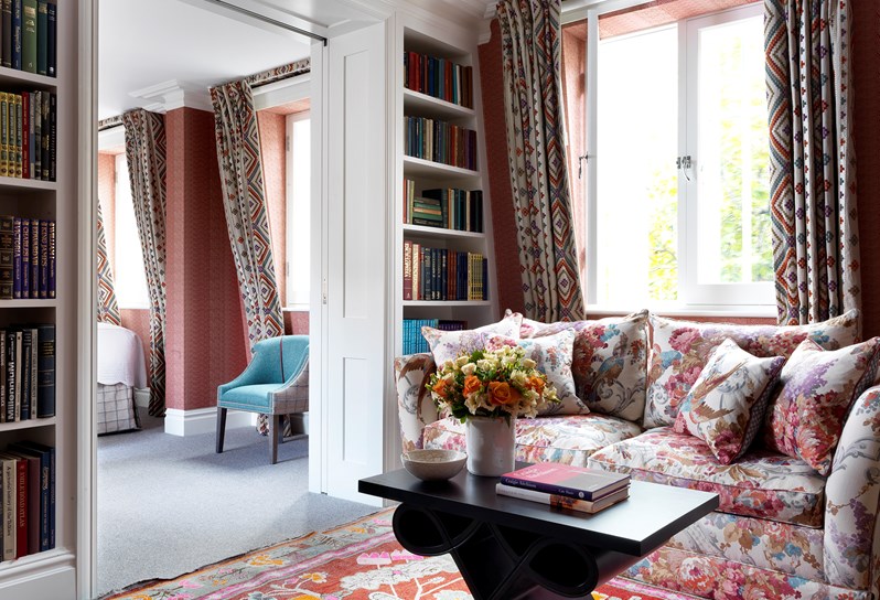 Living room of the Terrace Suite at Covent Garden Hotel with a flower pattern sofa and coffee table sitting over a patterned rug. To the left there is 2 bookcases with books on the wall, the door leading to the bedroom in between, where there is a chair and the corner of the bed