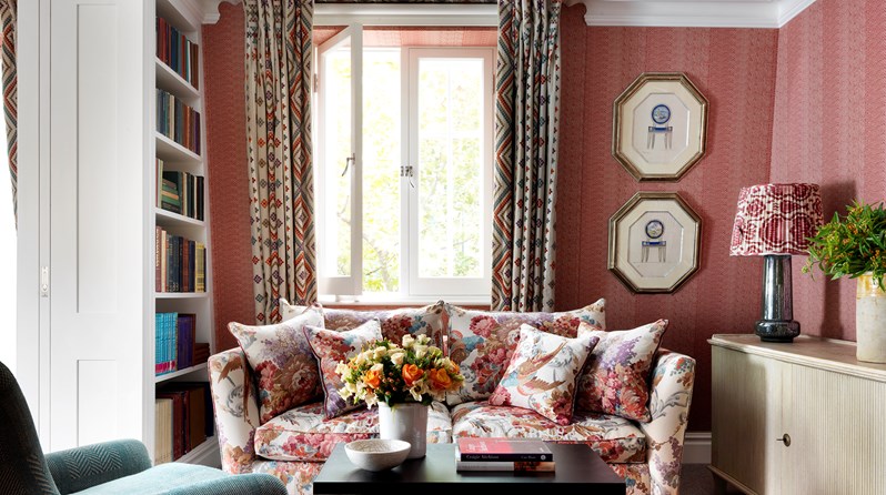 Living room of the Terrace Suite at Covent Garden Hotel with a flower pattern sofa and coffee table sitting over a patterned rug, there is a comfy arm chair opposite the sofa, on the left there is a bookcase and on the right a console with a lamp and flower arrangement over it.