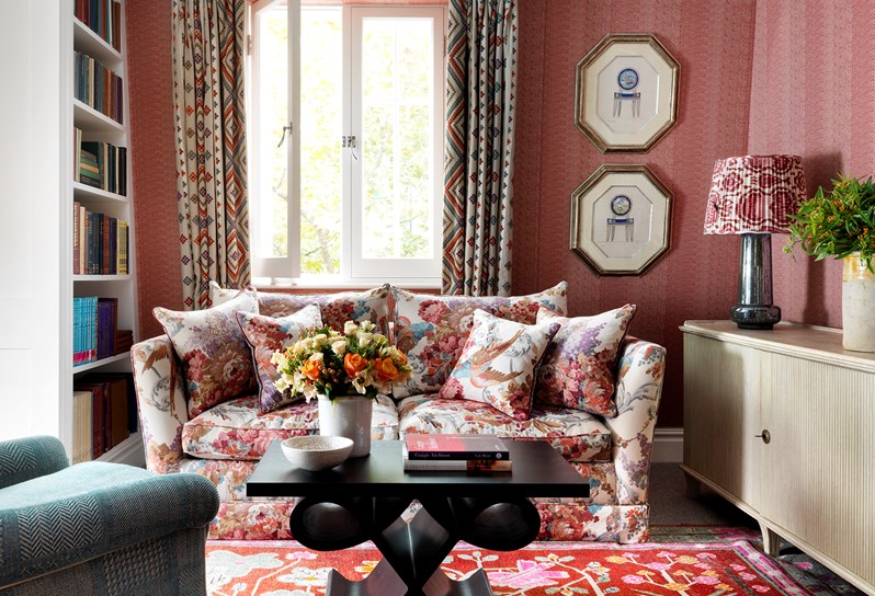 Living room of the Terrace Suite at Covent Garden Hotel with a flower pattern sofa and coffee table sitting over a patterned rug, there is a comfy arm chair opposite the sofa, on the left there is a bookcase and on the right a console with a lamp and flower arrangement over it.