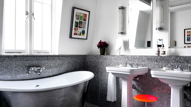 Grey granite bathroom with a silver free standing bathtub under the window, double vanity to the right side with a pink stool sitting between the two sinks.