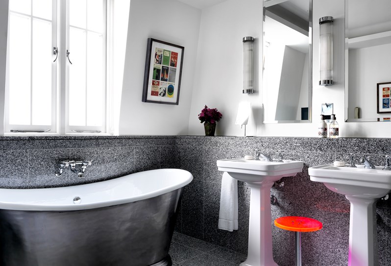 Grey granite bathroom with a silver free standing bathtub under the window, double vanity to the right side with a pink stool sitting between the two sinks.