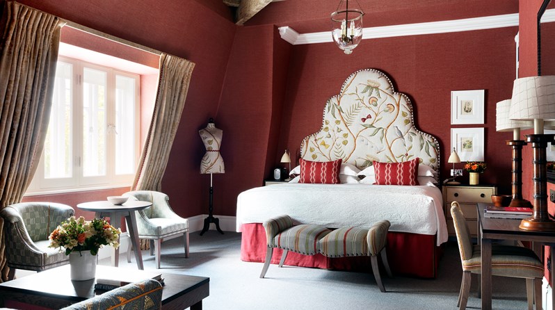 Bedroom of the Terrace Suite at Covent Garden Hotel. The bed sits at the back of the room with a pattern headboard, work space with desk and chairs to the right side of the bed. Two chairs and small table sits under the window on the left side. The room also has a sitting area with a sofa and coffee table opposite the bed.