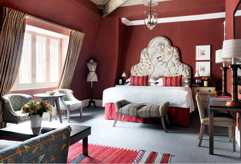 Bedroom of the Terrace Suite at Covent Garden Hotel. The bed sits at the back of the room with a pattern headboard, work space with desk and chairs to the right side of the bed. Two chairs and small table sits under the window on the left side. The room also has a sitting area with a sofa and coffee table opposite the bed.