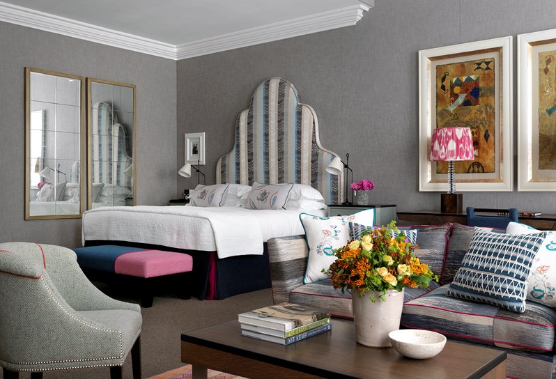 The living space of an open plan suite, with the corner of the bed on the left hand side, a desk with two lamps on, a side table and comfortable armchair in front of floor to ceiling windows and parts of a grey sofa on the right.