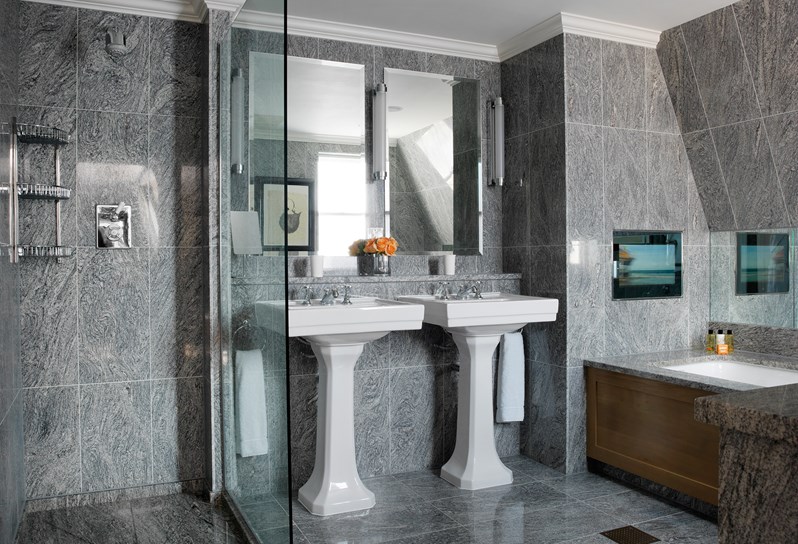 A beautiful granite bathroom with walk in shower to the left, two wash basins in the center and bathtub and television to the right