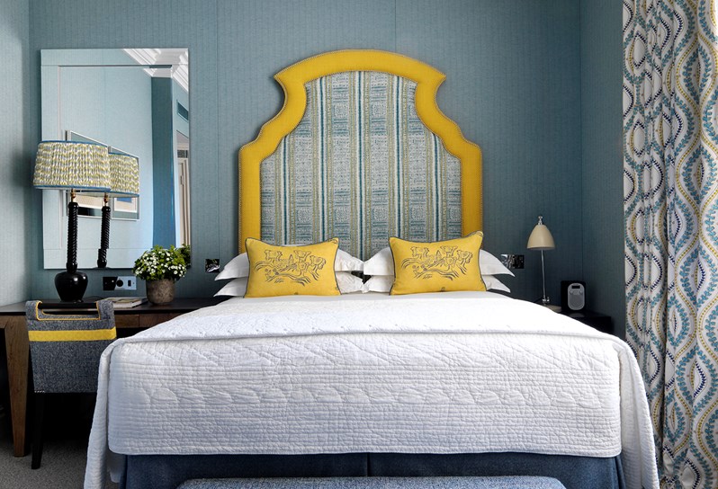Vertically striped grey and yellow wallpaper surrounds this beautiful bedroom, with a bright orange headboard and yellow cushions on the bed. There is a desk to the left and a grey and yellow ottoman at the end of the bed