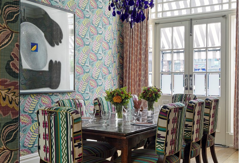 The Dora Carrington Room setup for a meeting. The far wall has a beautiful blue, lime green and pink wallpaper and there is a blue chandelier hanging above the table. At the end of the room there are doors that open onto Charlotte Street.