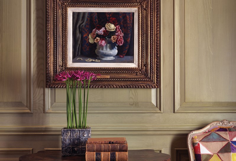 A beautifully framed painting of a vase of roses hangs on a wood paneled wall. There is a table below with antique books and plant with pink flowers and a colourful chair to the right of this.