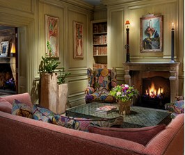 The Library with shelves of books either side of the fireplace. A comfortable red sofa and 2 colourful armchairs sit centered around a coffee table with fresh flowers.