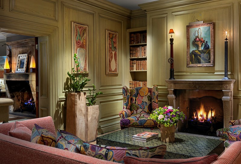 The Library with shelves of books either side of the fireplace. A comfortable red sofa and 2 colourful armchairs sit centered around a coffee table with fresh flowers.