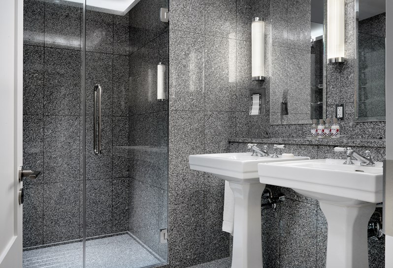A granite bathroom featuring a spacious walk in shower and two separate white porcelain basins with large mirrors hung above each one.