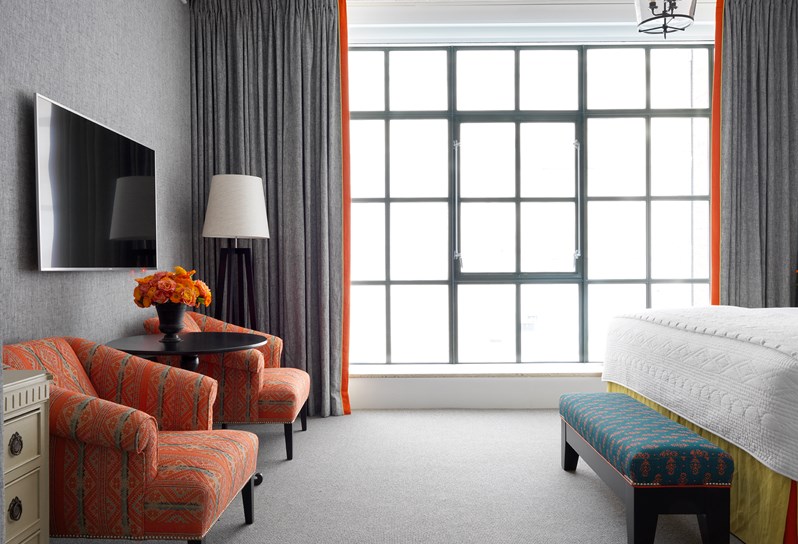 The seating area of a grey and orange bedroom with floor to ceiling windows, containing a side table with fresh color matched flowers between two comfortable arm chairs opposite the end of bed ottoman.