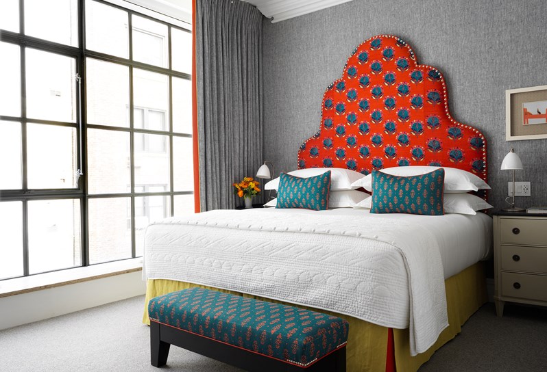 A king sized bed with a bold and colorful head board in a grey scaled bedroom and matching end of bed ottoman, with bright floor to ceiling windows.