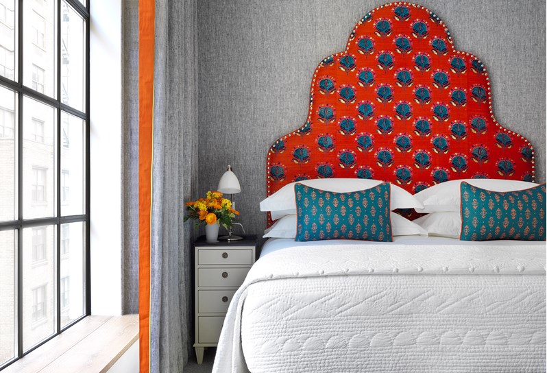 A close up of a comfortable king sized bed with a bright head board complementing the grey walls, next to floor to ceiling windows and a nightstand table with fresh flowers.