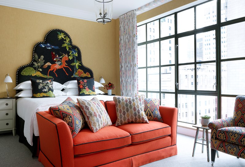 A bedroom with a king sized bed and floor to ceiling windows with views of Midtown Manhattan, with a playful, storytelling head board and a comfortable sofa and arm chair.