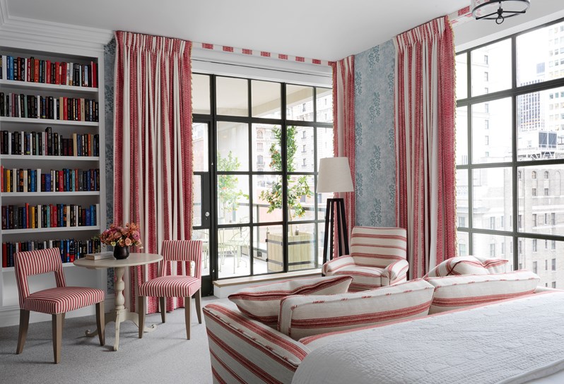 View from bed overlooking the corner of the room with floor to ceiling windows on two walls, showing a patterned blue wallpaper and floor to ceiling book case, contrasted by pinks and reds in striped curtains, two chairs, a sofa and a comfortable armchair.