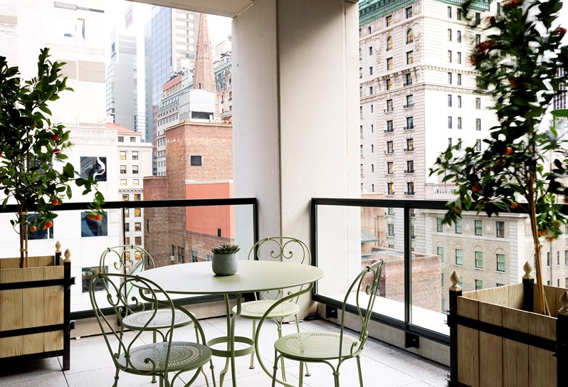 Corner terrace overlooking Midtown Manhattan, containing four metal summer green chairs and an accompanying table, surrounded by leafy plants.
