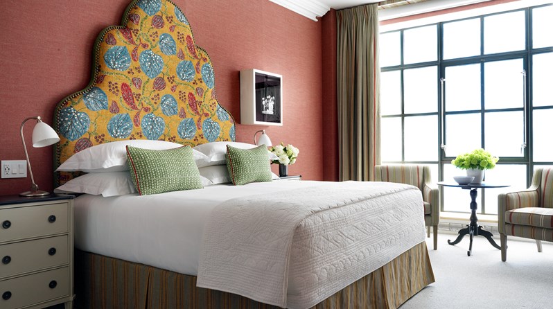 A spacious bedroom with colorful walling with the king sized bed boasting a playful headboard in yellow, blue and red tones. A side table and two striped arm chairs are placed in front of the floor to ceiling windows.