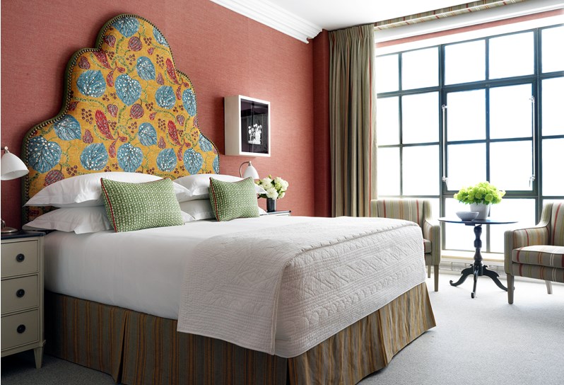 A spacious bedroom with colorful walling with the king sized bed boasting a playful headboard in yellow, blue and red tones. A side table and two striped arm chairs are placed in front of the floor to ceiling windows.