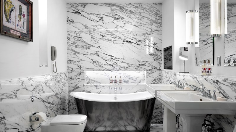 A spacious white marble bathroom featuring a free standing chrome bathtub, two separate basins, a toilet and a bidet, with Kit Kemp's own bathroom range, RikRak, placed throughout.