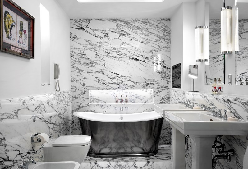 A spacious white marble bathroom featuring a free standing chrome bathtub, two separate basins, a toilet and a bidet, with Kit Kemp's own bathroom range, RikRak, placed throughout.