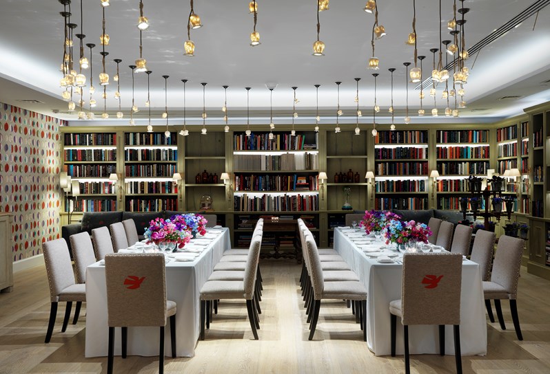 The Reading Room set for private dining for 24 people on two long tables, complete with comfortable chairs, beautiful table settings and colorful flower arrangements as centerpieces, in the surroundings of book lined walls and hanging lights in a circular array.