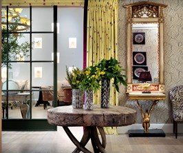 The entry to the Drawing Room with playful wallpaper and a large mirror, fronted by a large, bespoke, wooden table with fresh flowers in three vases on top.