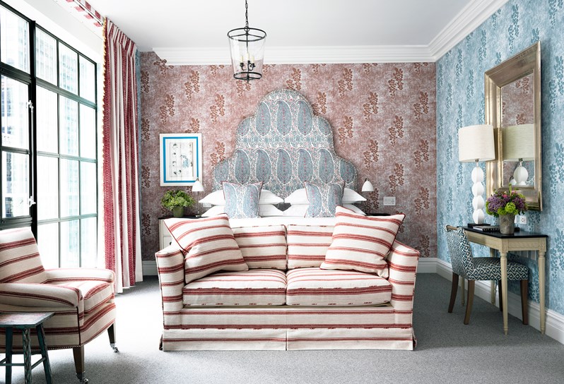 A bedroom with floor to ceiling windows, contrasting red and blue patterned wallpaper, a comfortable king sized bed with matching head board, a desk and chair and a comfortable sofa and arm chair with striped fabric.