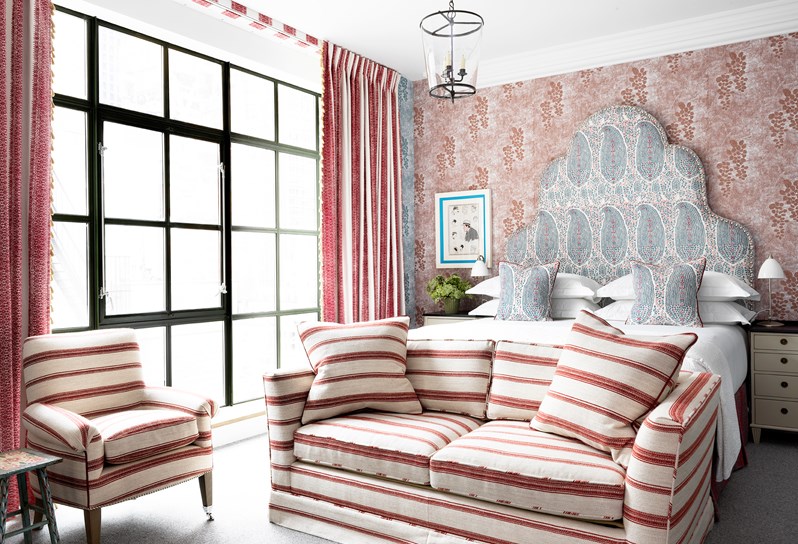 A bedroom with floor to ceiling windows, showing an inviting king sized bed with a statement headboard in red and blue, with contrasting red and blue patterned wallpaper. Featuring a comfortable sofa and arm chair with stripes.