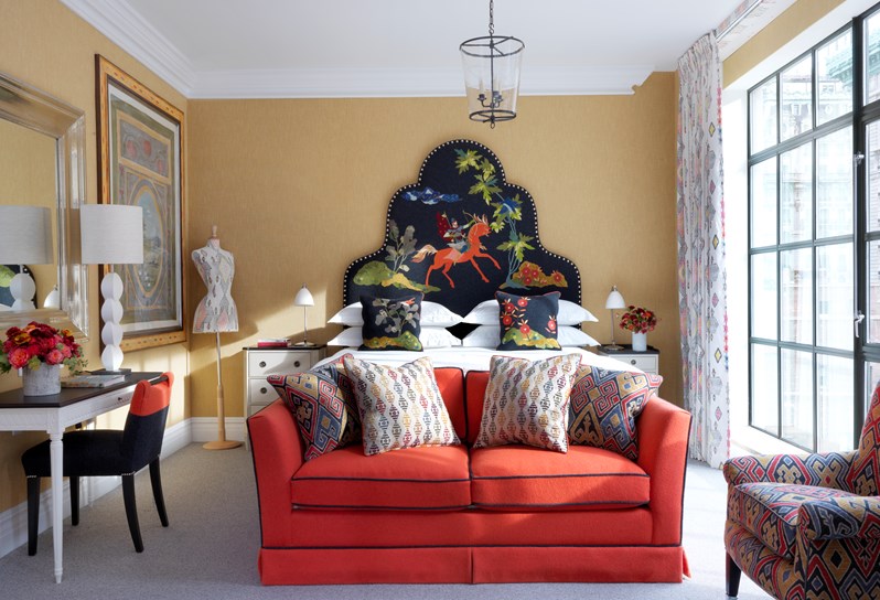 A light bedroom featuring a king sized bed with a statement head board, a desk and chair, a colorful sofa and armchair and Kit Kemp's statement mannequin in matching pattern to curtains and pillows.