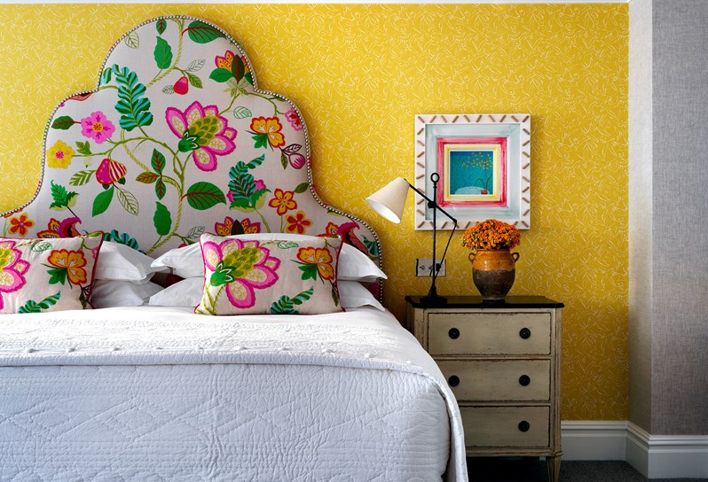 A bright yellow wall with a colorful picture hung above a nightstand table containing a lamp and fresh flowers, placed next to a large bed with a flowery headboard in multiple, bright colors.
