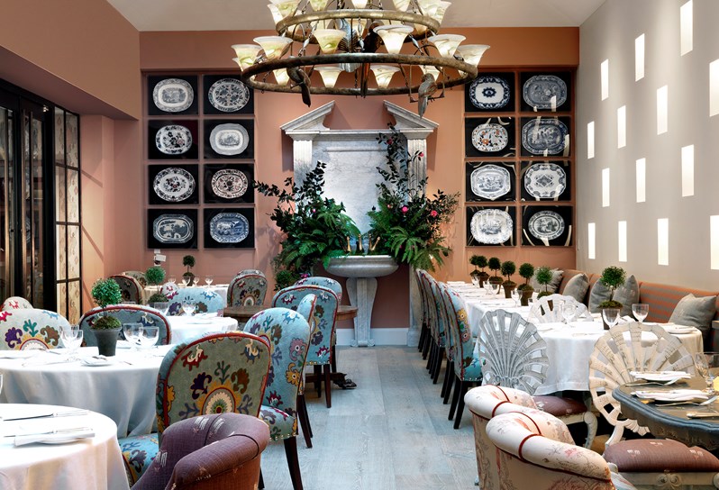 The Orangery with plenty of comfortable seats and tables for dining. A wall with sixteen art pieces showcasing various serving plates scattered around a leafy fountain oasis.
