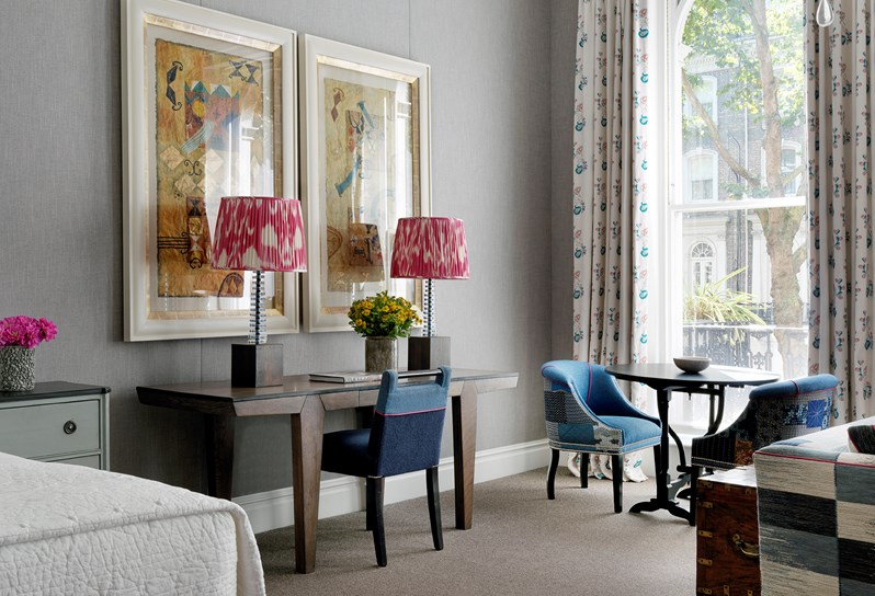 A view from a seating area featuring a comfortable sofa and armchair around a coffee table with fresh flowers, looking towards a bed with a large headboard in blue tones and an end of bed foot stool in matching colors.