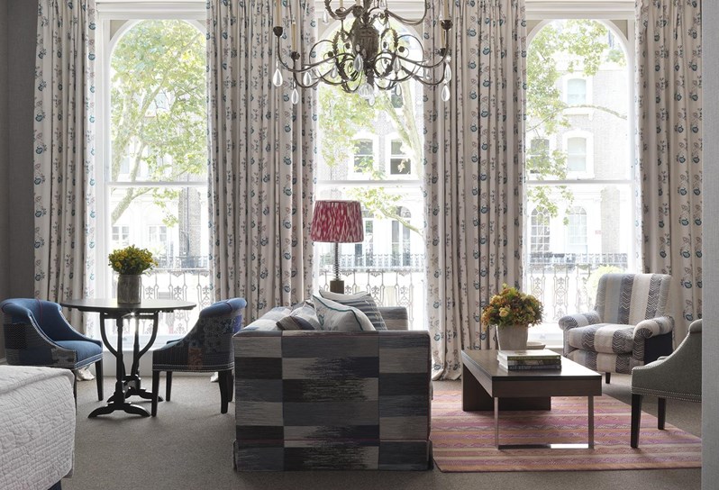 The Knightsbridge Suite at Knightsbridge Hotel. A view straight towards three floor to ceiling windows, fronted by a side table between two armchairs, and a seating area with a large grey sofa and a bright pink armchair arranged around a coffee table with fresh flowers in matching colors.