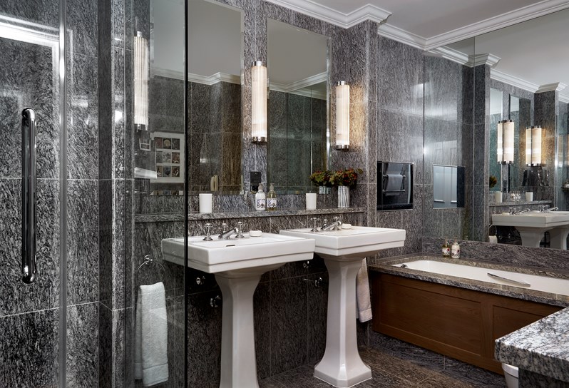 A closeup of a granite bathroom, featuring a walk in shower, two pedestal basins under large mirrors and a deep bathtub with a TV in.