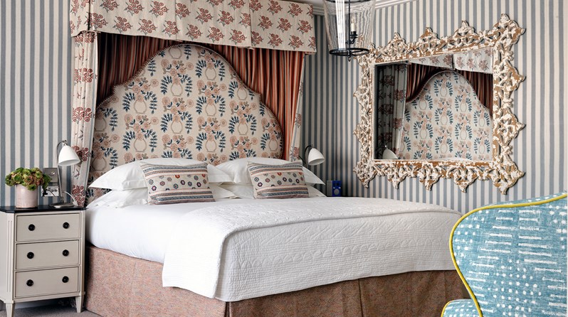 A spacious bedroom with soft, striped walls, boasting a king sized bed with a half tester and large, floral headboard