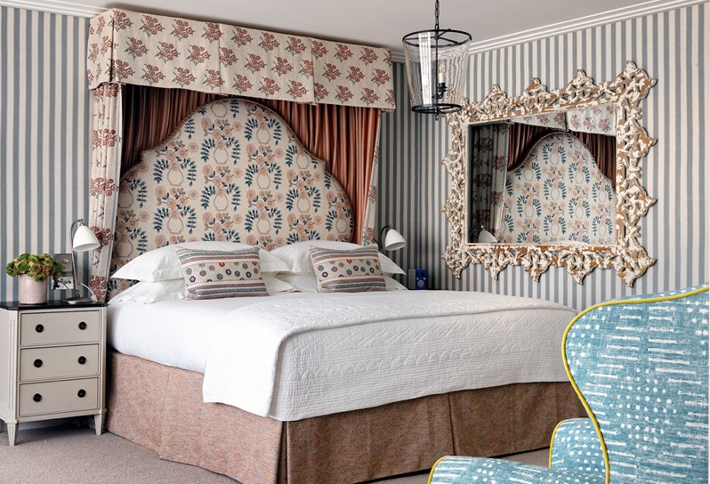 A spacious bedroom with soft, striped walls, boasting a king sized bed with a half tester and large, floral headboard