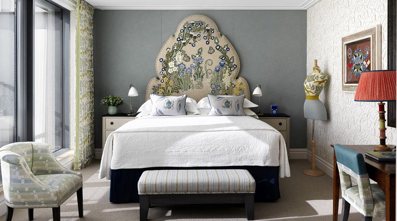Ham Yard Hotel's Terrace Suite bedroom. A king sized bed with a colourful, floral embroidered headboard, centered in a blue grey bedroom with cream nightstand tables on both sides, as well as a striped footstool at the end of the bed.