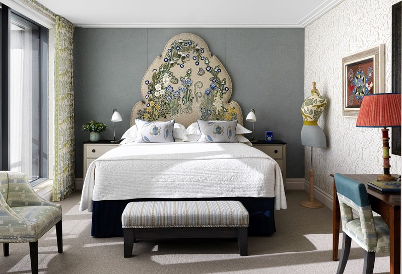 Ham Yard Hotel's Terrace Suite bedroom. A king sized bed with a colourful, floral embroidered headboard, centered in a blue grey bedroom with cream nightstand tables on both sides, as well as a striped footstool at the end of the bed.