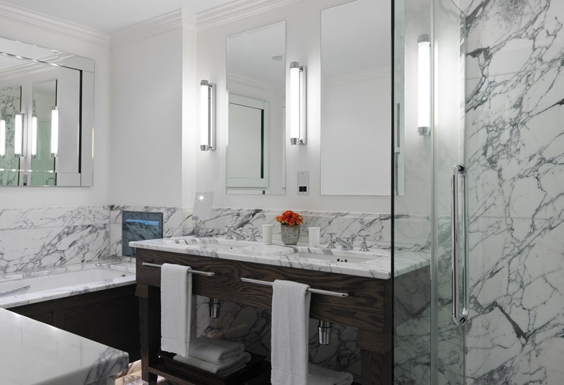 A spacious bathroom in white marble and wooden details, with a deep bath containing a TV, a large walk in shower, two separate sinks and several mirrors throughout the room.