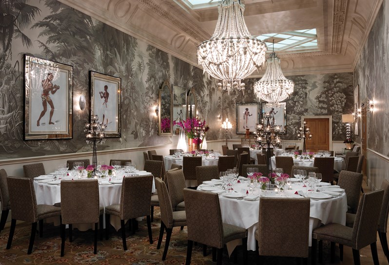 The Shooting Gallery at Haymarket Hotel with dramatic high ceilings, ornate cream chandeliers and graphic grey wallpaper with forest mural. Several round white tables are set for a formal dinner with beautiful pink floral arrangements on the centre of the table.